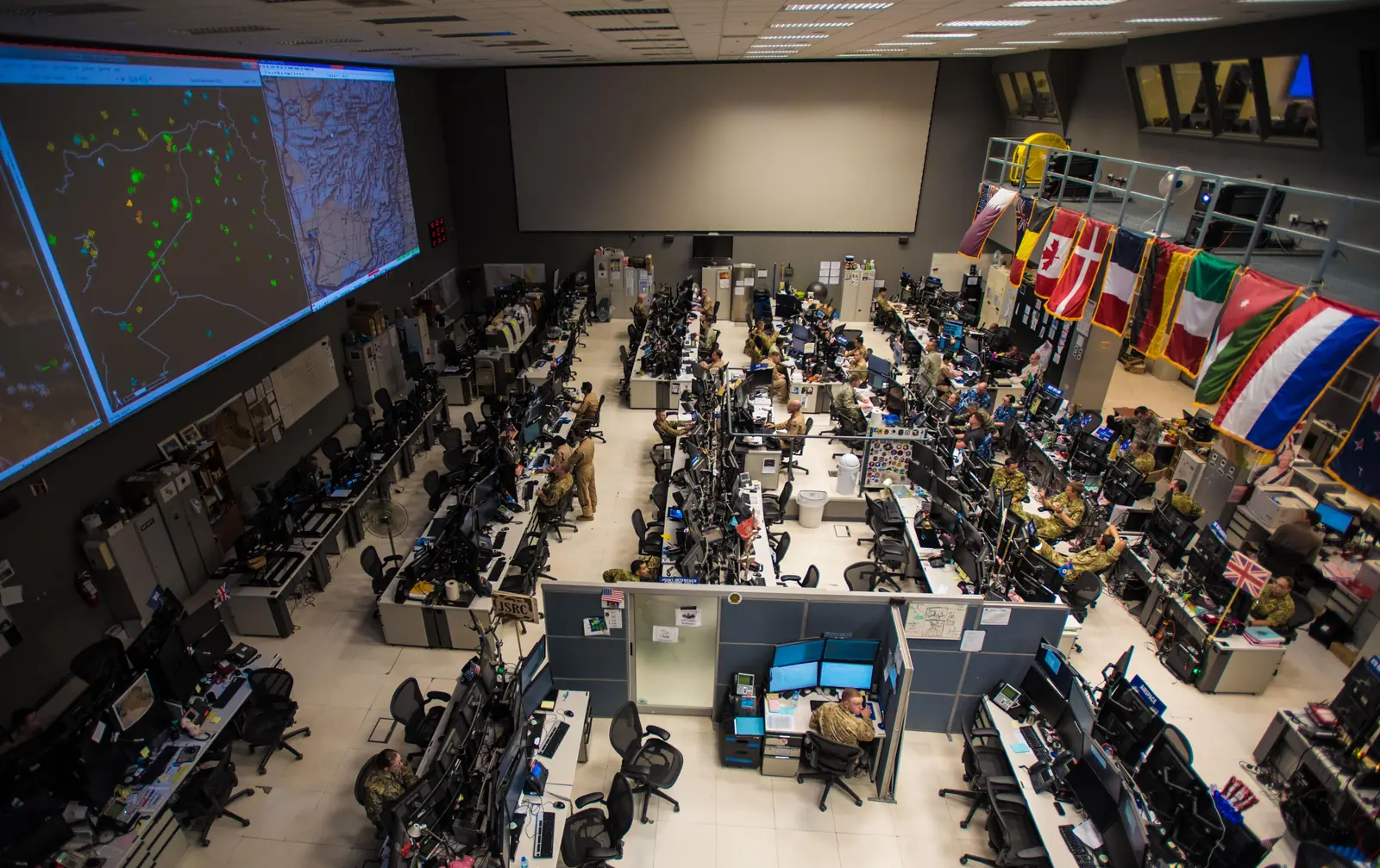 A large military operations room with rows of uniformed personnel at computer workstations facing wall-sized screens and a row of national flags