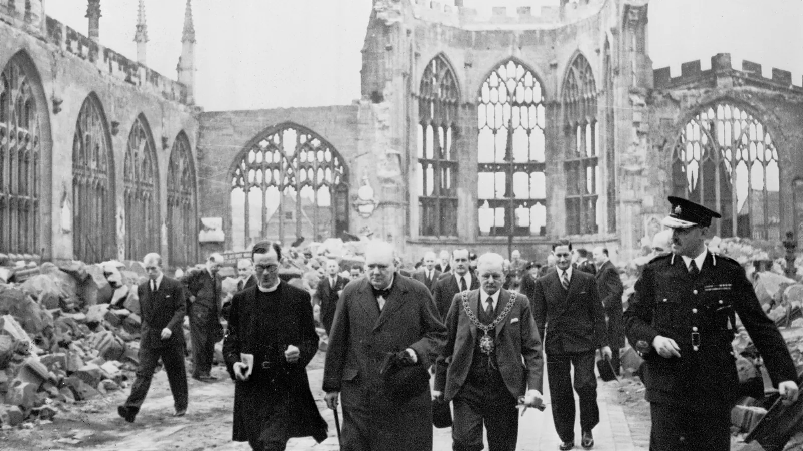Winston Churchill walks through the bombed-out ruins of Coventry Cathedral with officials, rubble piled on either side