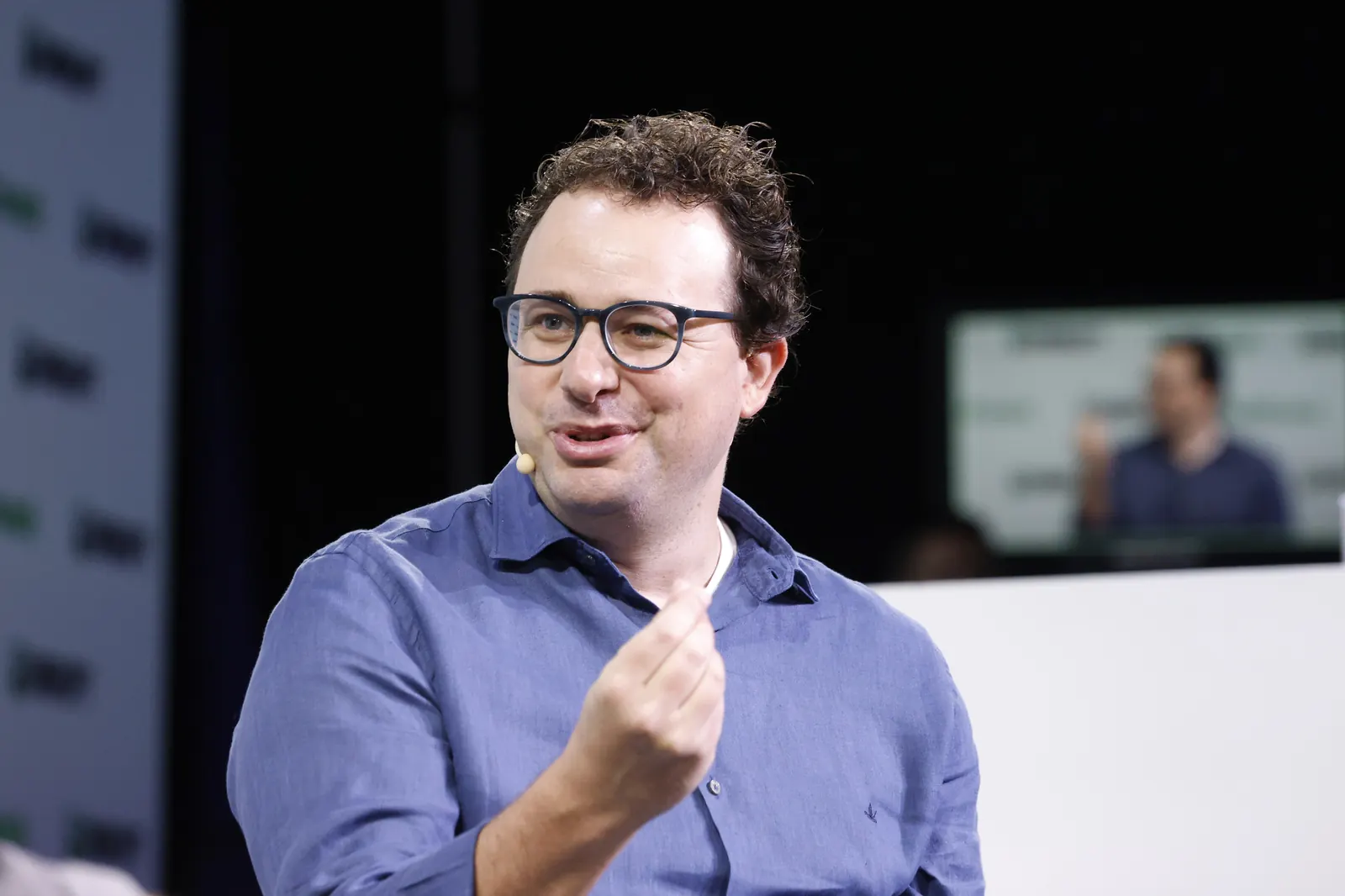 Dario Amodei speaking on stage at a technology conference, gesturing with one hand, wearing glasses and a blue button-down shirt
