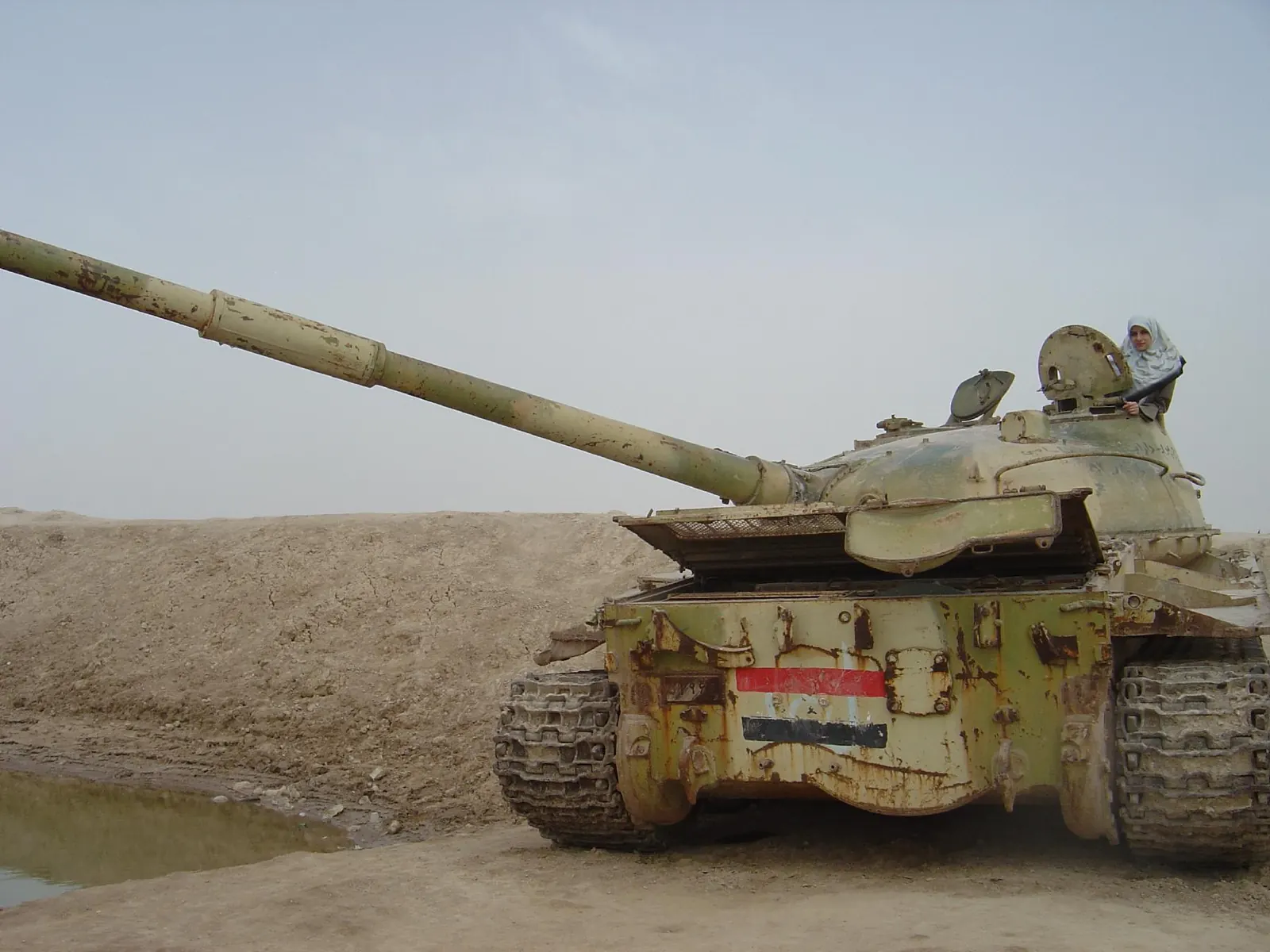 A disabled Iraqi T-62 tank with the Iraqi flag painted on its hull, sitting in desert sand behind an earthen berm near the Iran-Iraq border