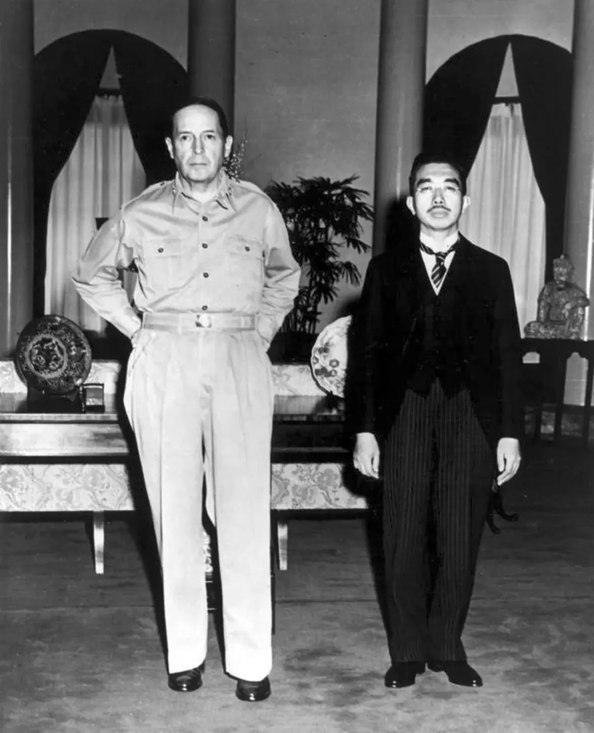 Emperor Hirohito in a dark suit standing next to General Douglas MacArthur in open-collar khaki uniform at the US Embassy in Tokyo