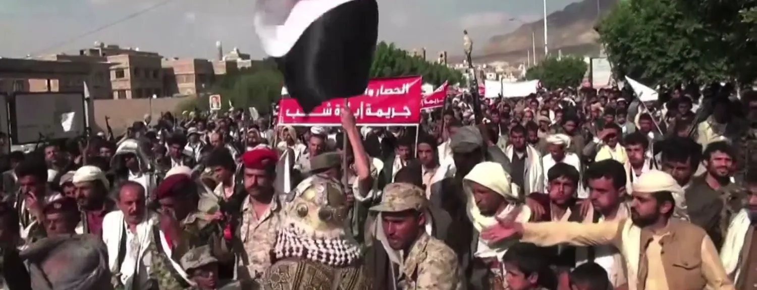 A Houthi rally in Sanaa, with armed men in the crowd, red banners, and a Yemeni flag waving overhead