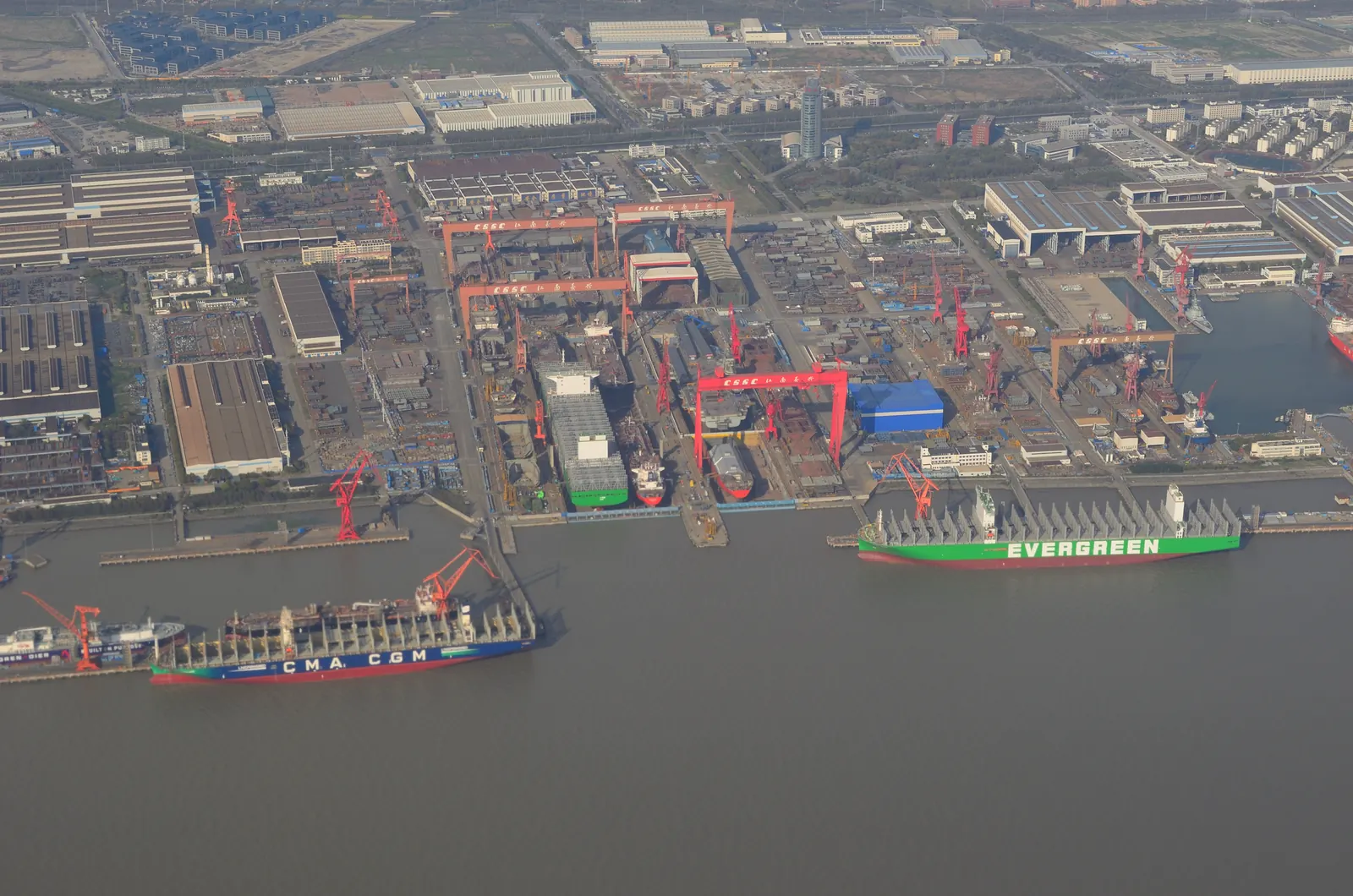 Aerial view of the Jiangnan Shipyard in Shanghai, with rows of red gantry cranes over covered building docks and two large ships fitting out at the quay