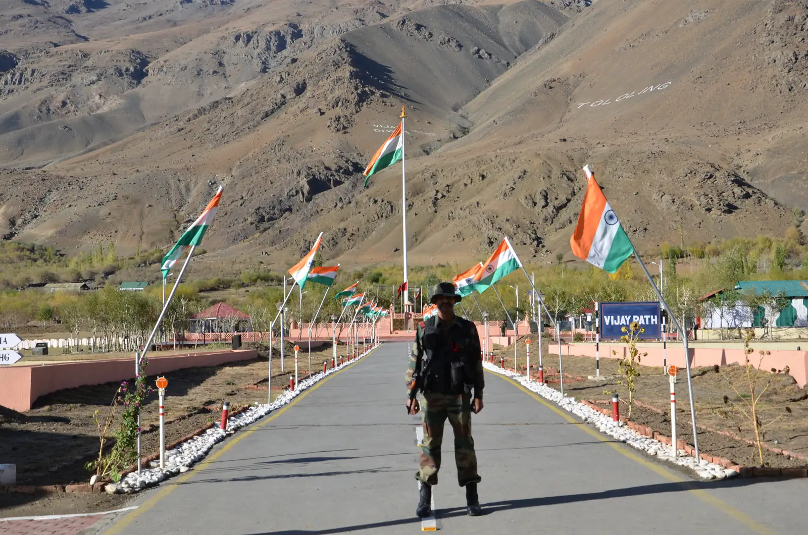 An armed Indian Army soldier on a flag-lined path at the Kargil War Memorial in Drass, with barren Himalayan war-zone peaks behind him