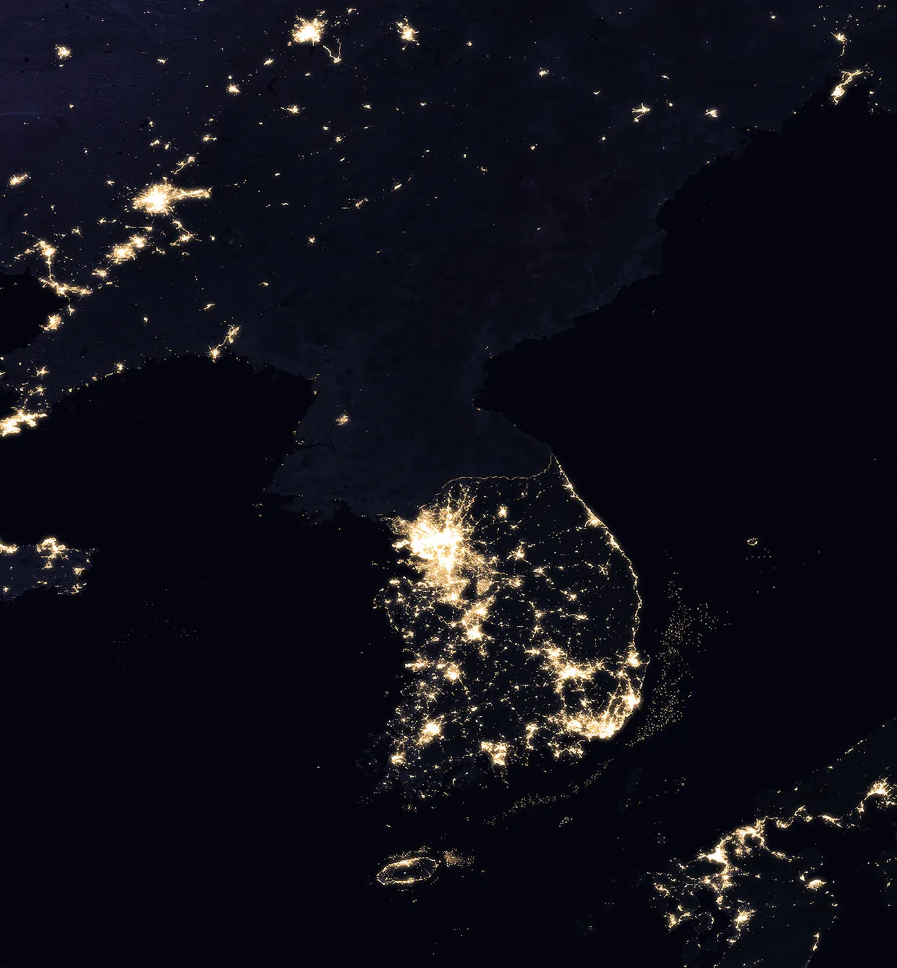 The Korean Peninsula at night as seen from space, with South Korea brightly lit and North Korea almost entirely dark except for a small glow around Pyongyang