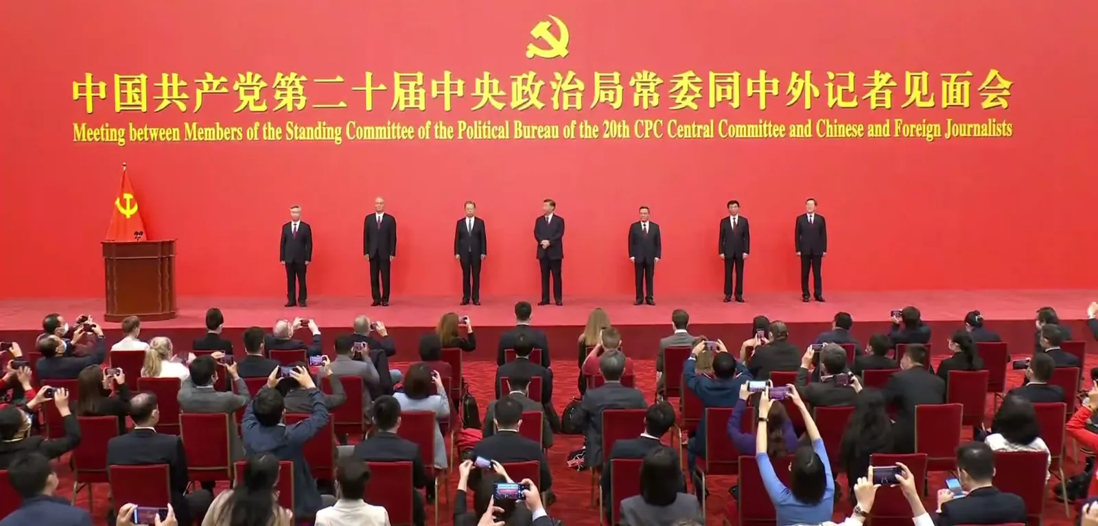 The seven members of the Politburo Standing Committee standing in a line on a red stage as rows of photographers raise their cameras
