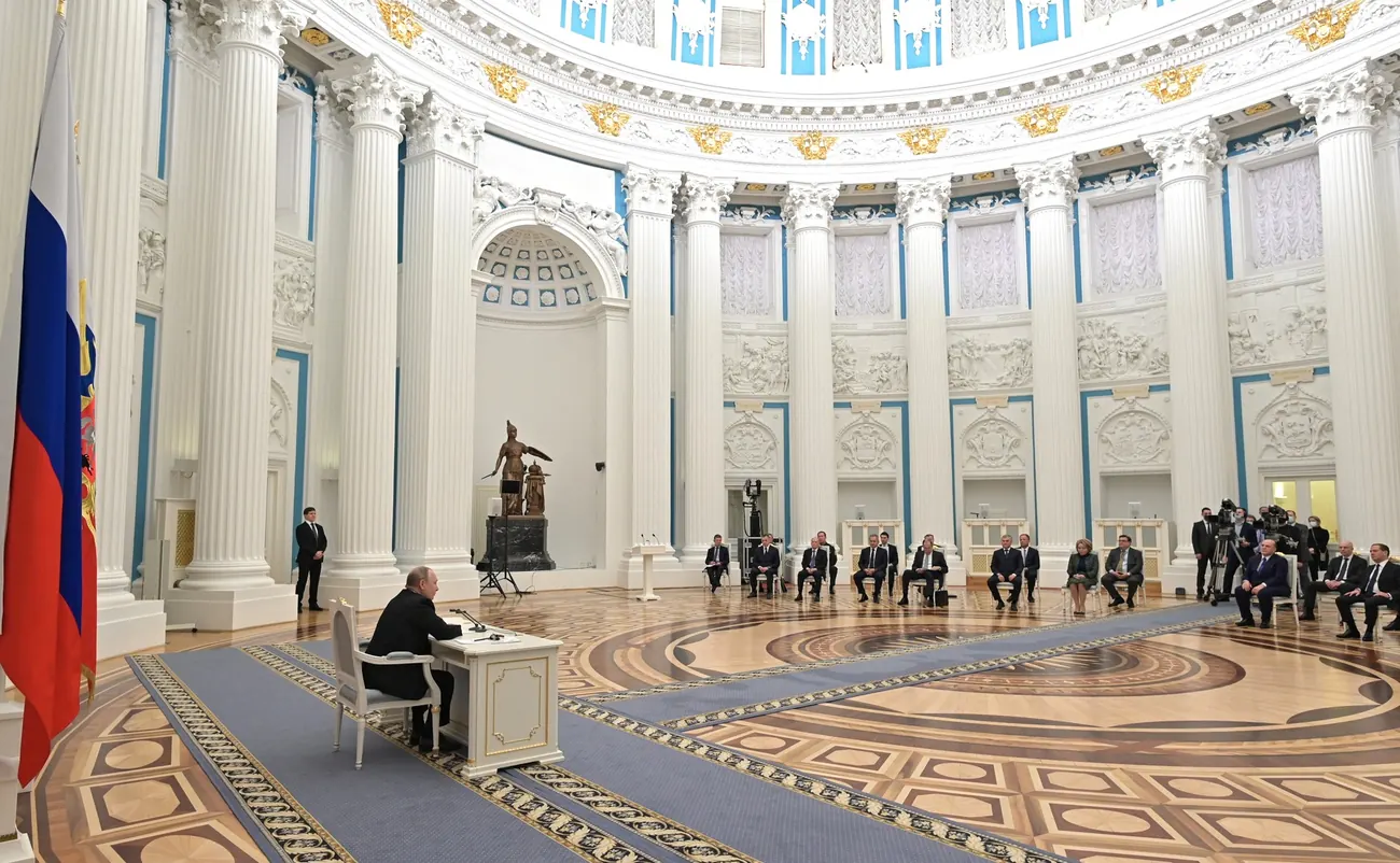 Vladimir Putin at a desk in the Kremlin, his Security Council members seated in chairs spread across the room