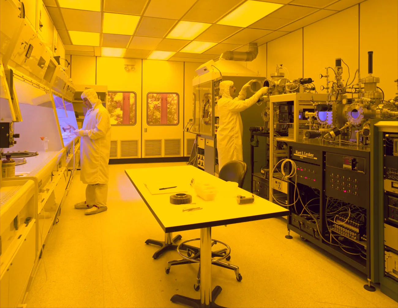 Two engineers in cleanroom suits working with semiconductor fabrication equipment under yellow light