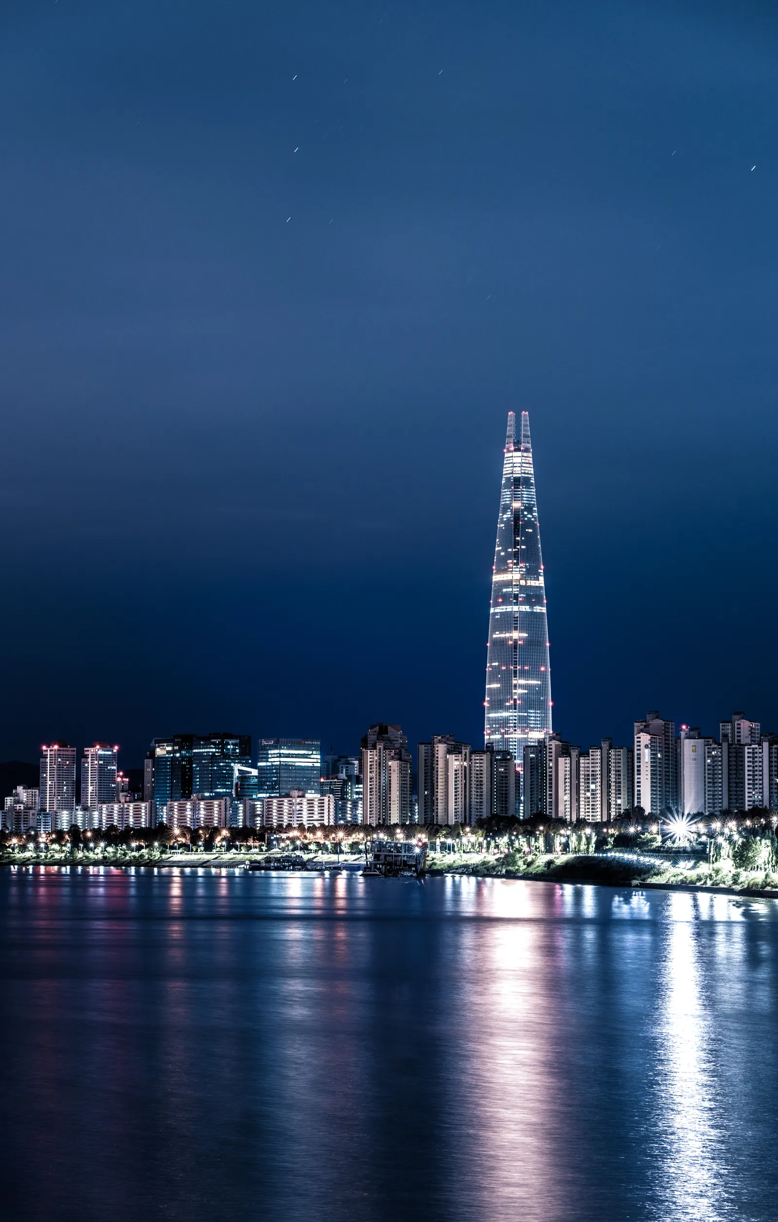 The Seoul skyline at night with the Lotte World Tower rising above illuminated buildings reflected in the Han River