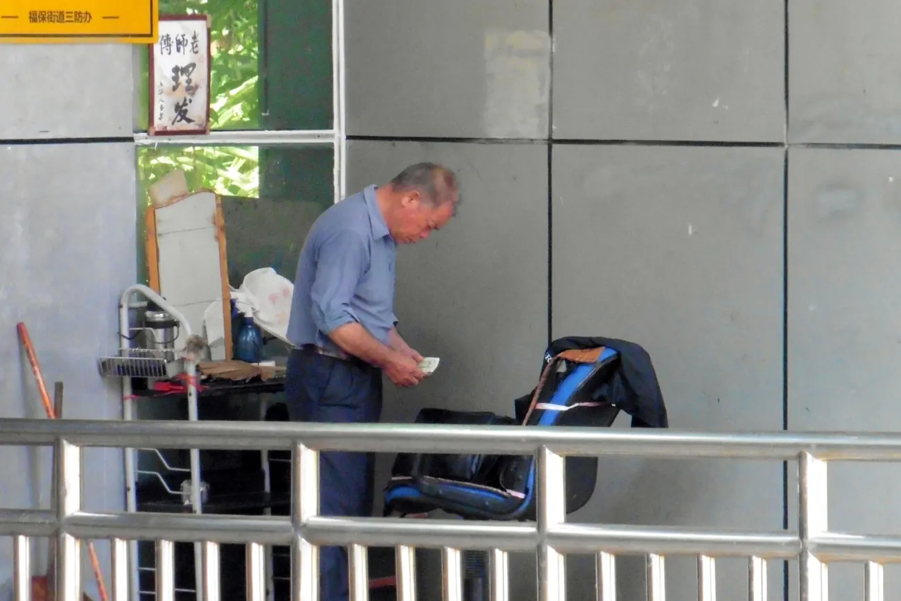 A street barber in Shenzhen counting a banknote beside his folding chair and mirror