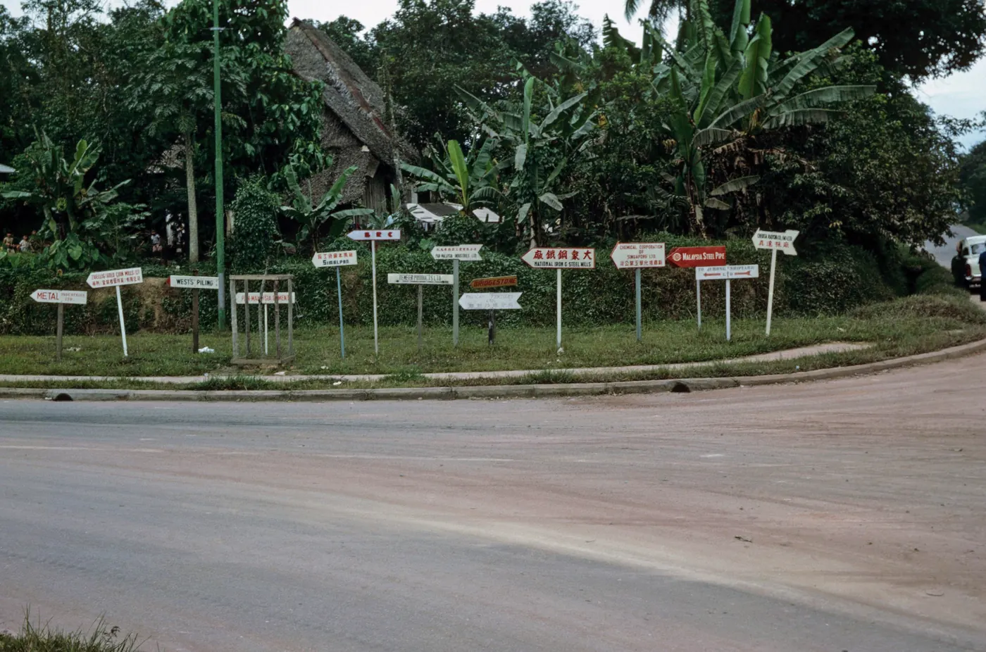 Signs pointing to factories at Jurong Industrial Estate in Singapore, June 1964, with tropical vegetation and a thatched-roof hut in the background