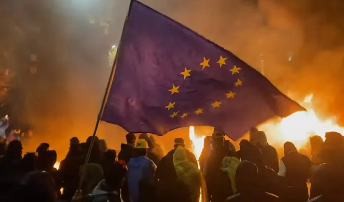 A European Union flag held above a crowd with burning barricades behind it at night