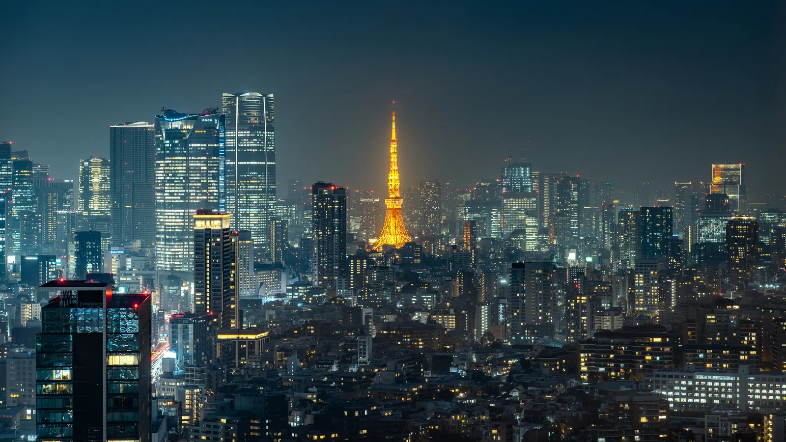 The Tokyo skyline at night with Tokyo Tower glowing orange at center, surrounded by dense high-rises and city lights