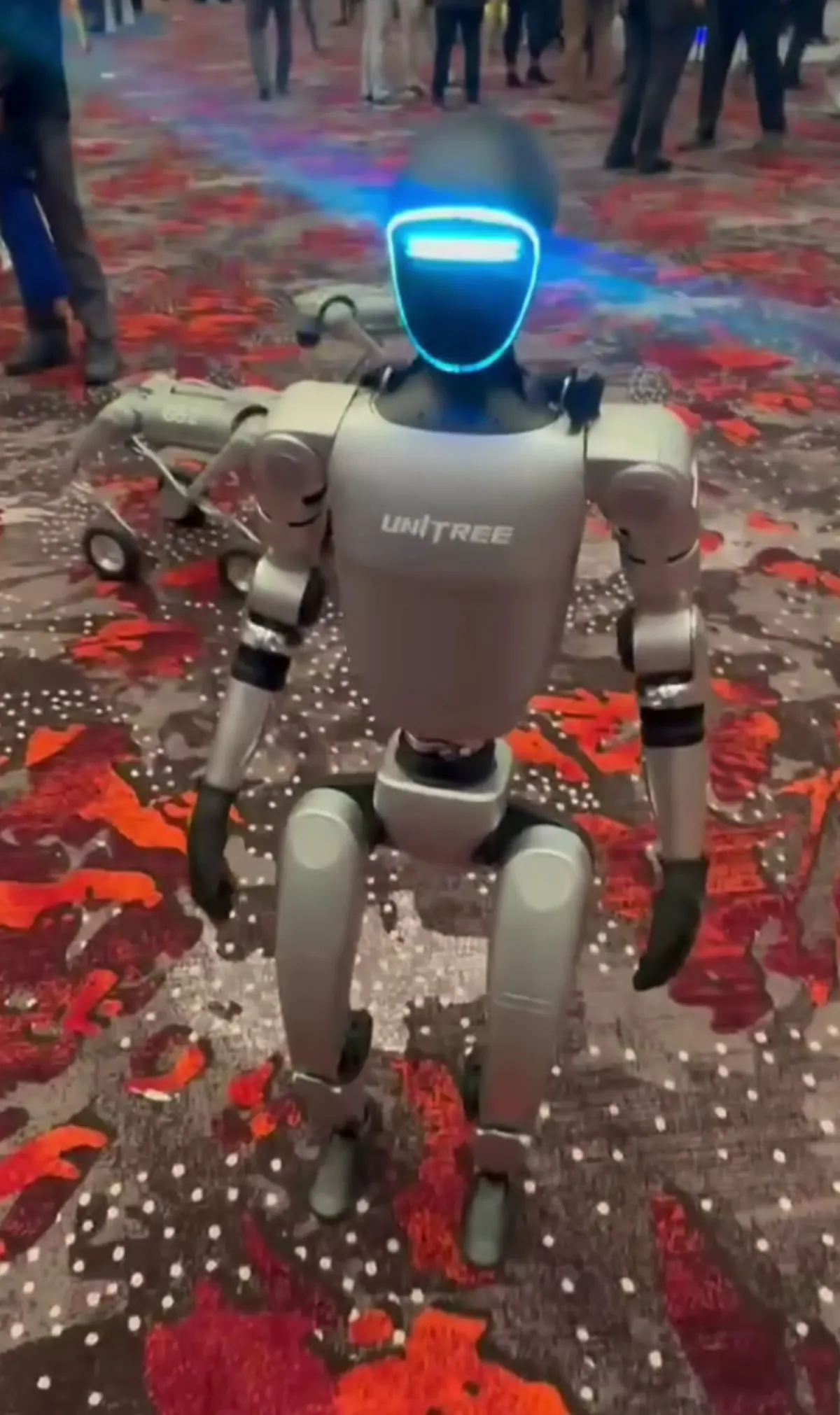 A Unitree G1 humanoid robot with a glowing blue visor, standing on a patterned carpet at a technology expo