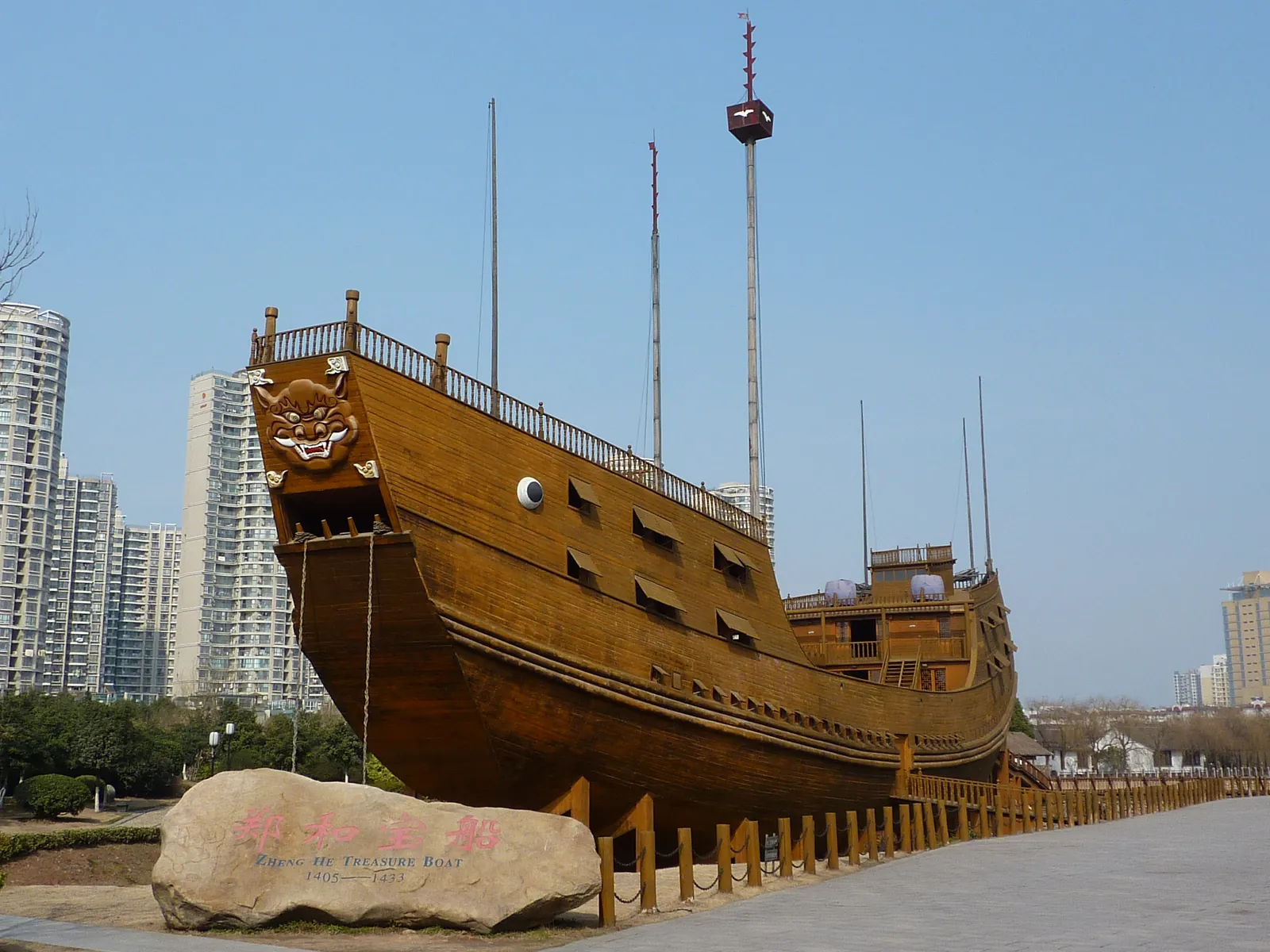 A full-size wooden replica of a Zheng He treasure ship in a park in Nanjing, with a stone marker reading Zheng He Treasure Boat 1405-1433 in the foreground and modern skyscrapers behind it