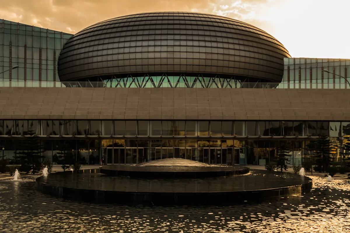 The domed conference hall of the African Union's headquarters in Addis Ababa, seen across a round pool with fountains under an orange evening sky