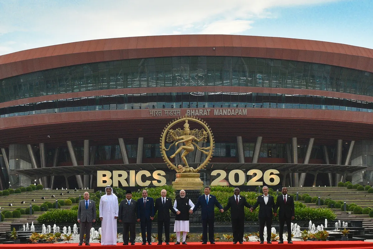 Ten leaders and envoys stand in a row on a red carpet in front of the Bharat Mandapam convention center in New Delhi, under large letters reading BRICS 2026