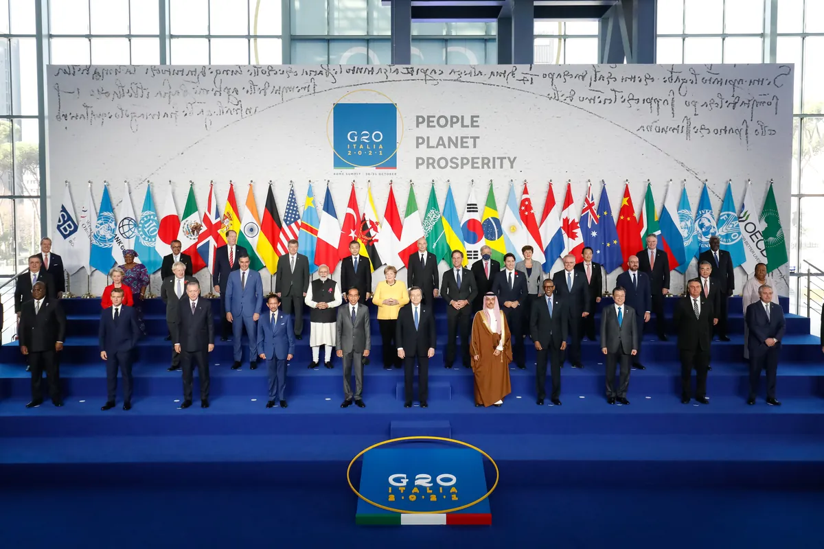 Leaders of the G20 and invited countries stand on blue steps in front of a row of national flags, under a sign reading G20 Italia 2021 and People, Planet, Prosperity