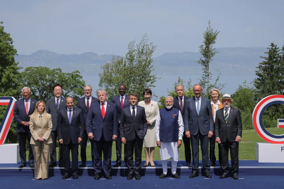 Leaders of the G7 and of invited countries stand in two rows on a blue stage between giant G7 letters, with Lake Geneva and mountains behind them