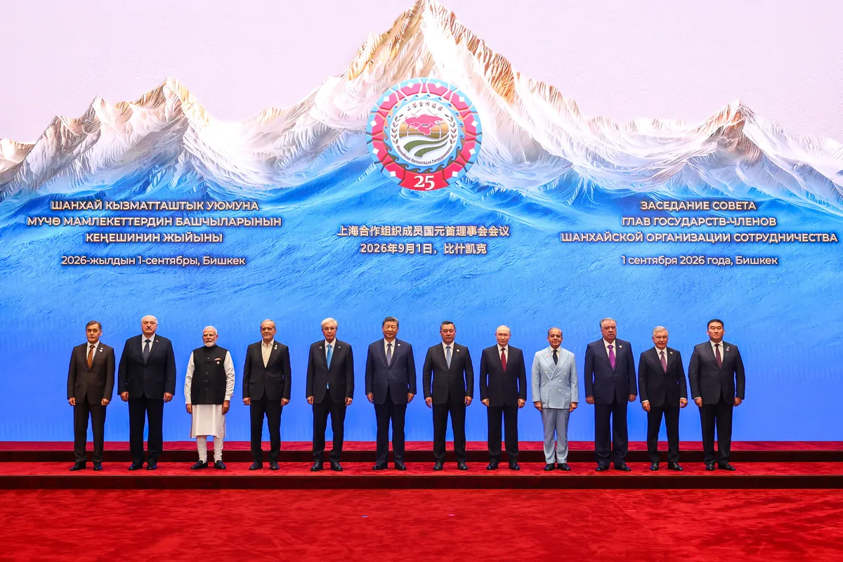 Twelve leaders stand in a row on a red carpet, in front of a painted backdrop of snowy mountains and the SCO's 25th anniversary emblem