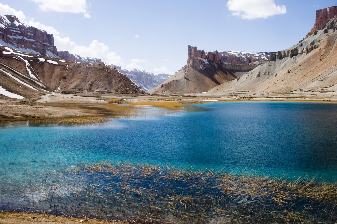 A turquoise lake in Band-e Amir National Park, with reeds in the foreground and snow-streaked cliffs behind