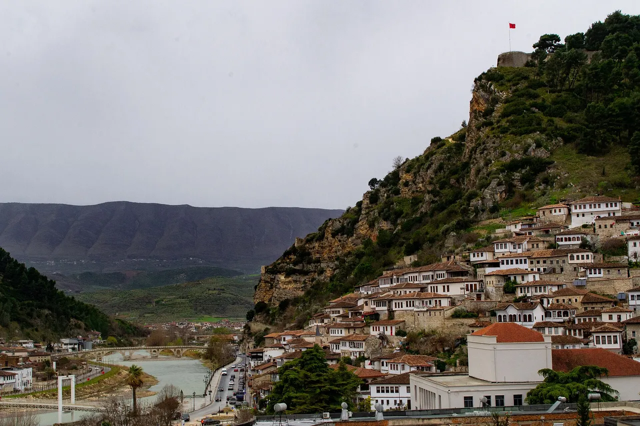 White Ottoman-era houses with rows of windows climbing a steep green hillside below a castle, with a river and mountains behind