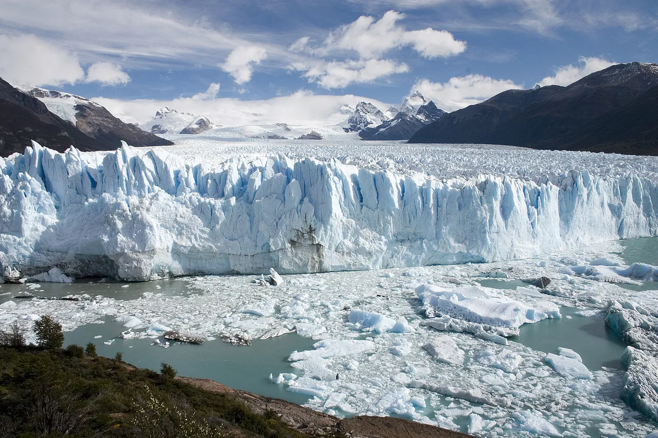 The ice wall of the Perito Moreno Glacier above a lake full of floating ice, with snowy peaks behind