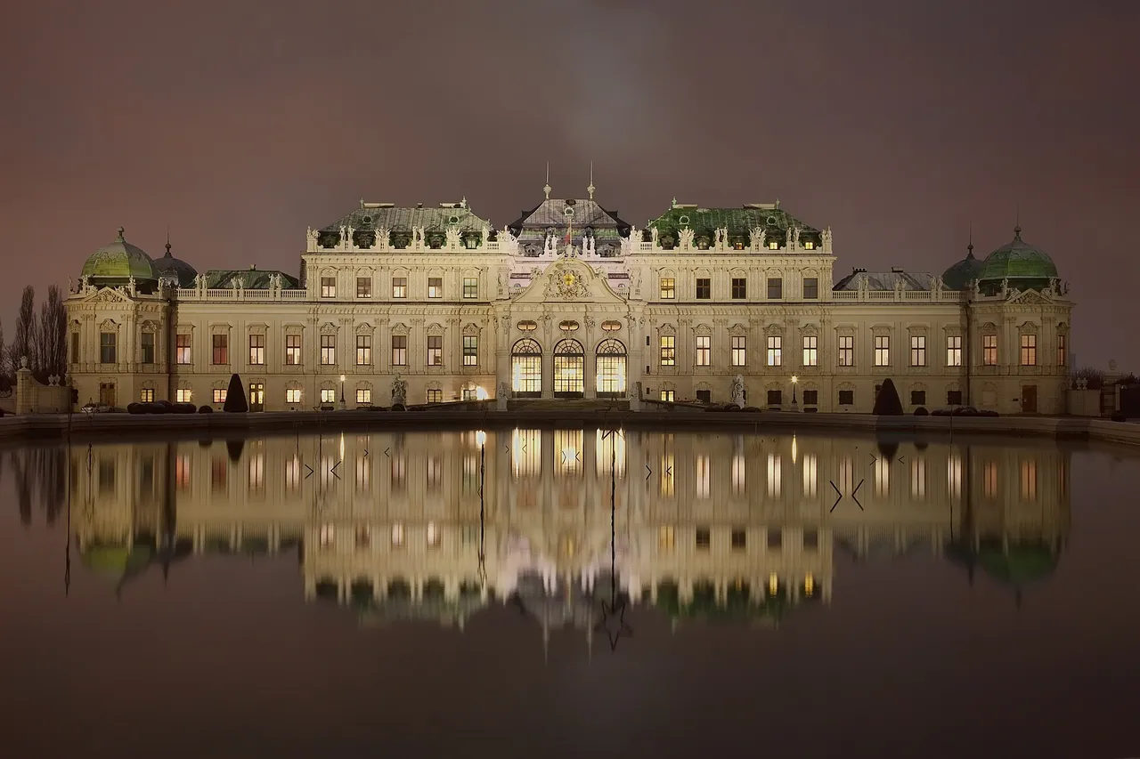 The Upper Belvedere palace in Vienna lit up at dusk, with its long baroque facade and green domes reflected in a still pond