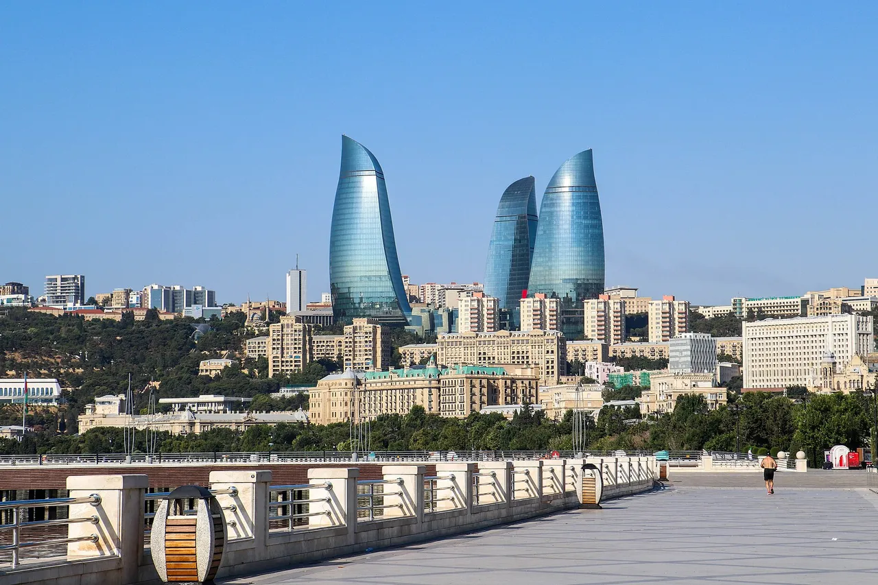 Three curved glass towers rise over the city behind a wide seaside promenade