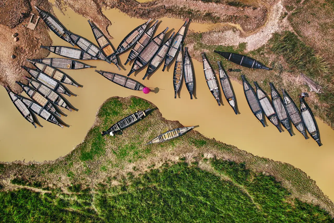 Aerial view of dozens of long wooden boats moored along the muddy banks of a river, with green grass on the islands between the channels