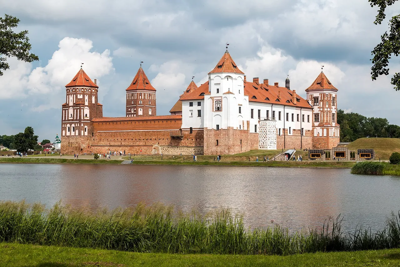 A red-brick castle with orange-roofed towers and a white central building, beside a pond under a cloudy summer sky