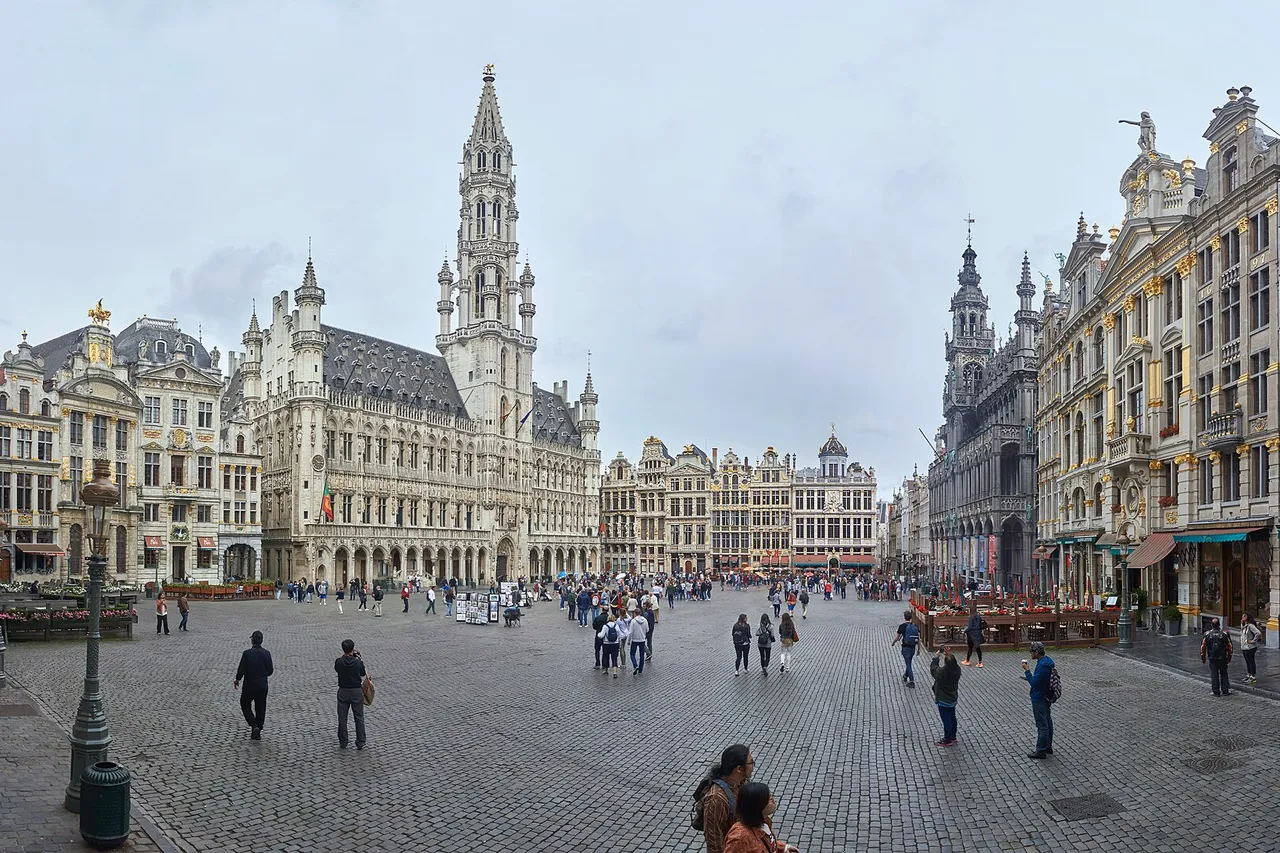The Gothic town hall with its tall spire and the ornate guildhouses around the cobbled Grand-Place in Brussels, with people walking across the square