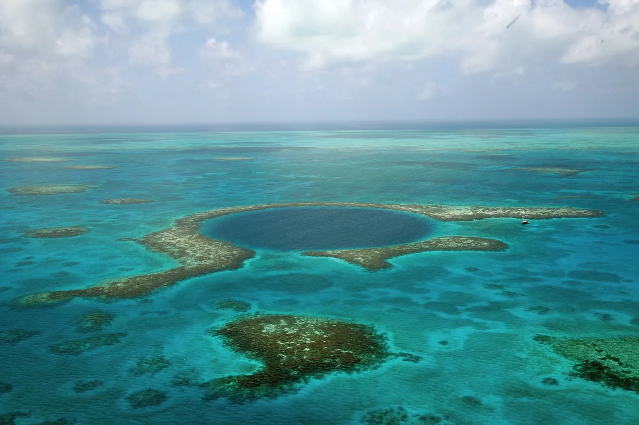 A dark blue round hole in the middle of a turquoise coral reef, seen from the air, with reef patches around it and clouds on the horizon