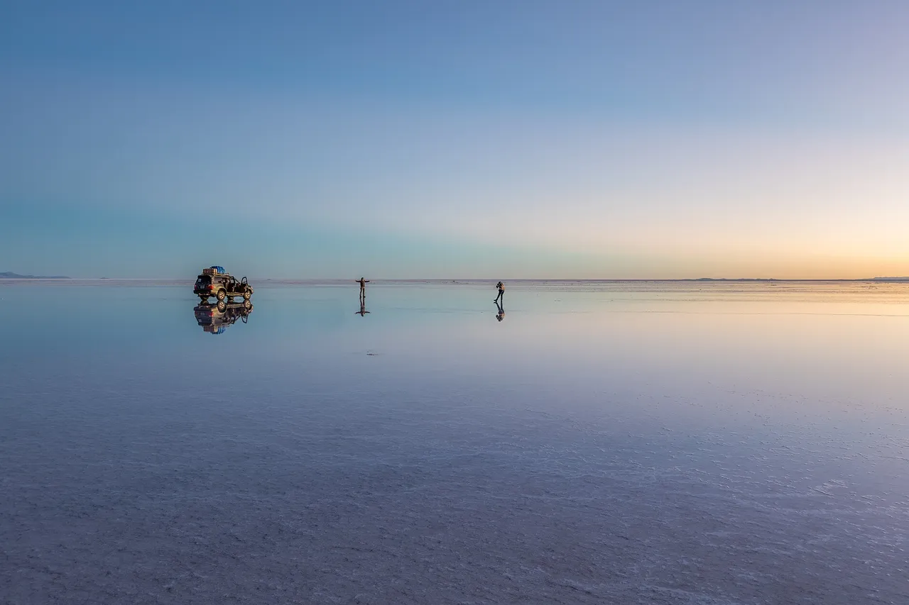 A thin layer of water on the Uyuni salt flat mirrors the dawn sky, with a jeep and two people far away