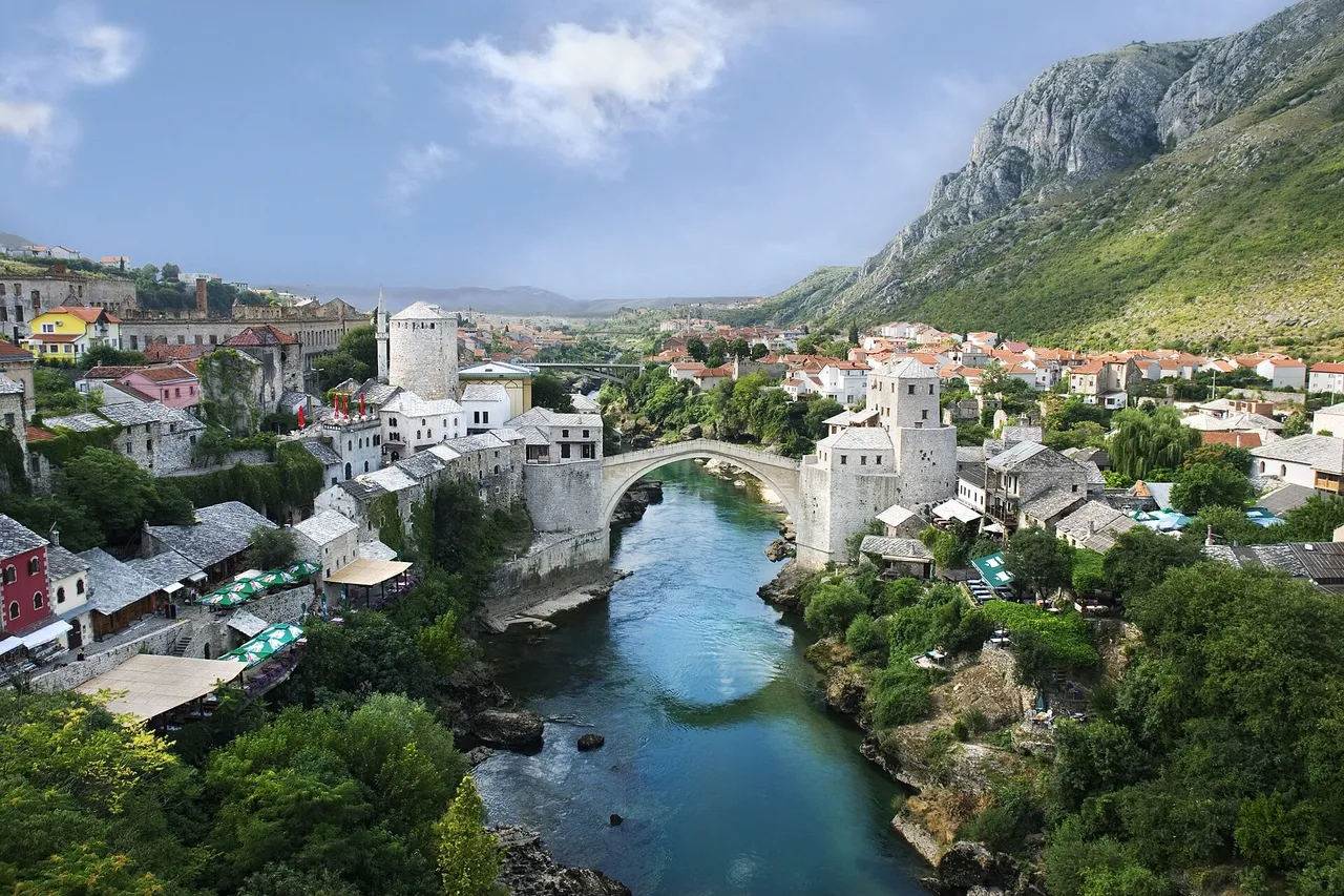 The Old Bridge in Mostar arching over the green Neretva river between stone towers, with old houses, trees and a steep mountainside around it
