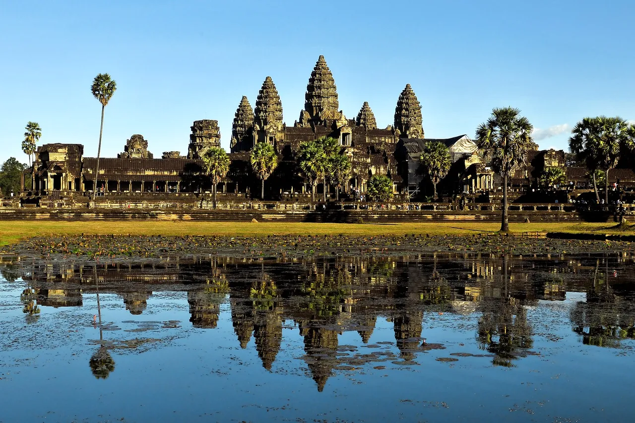 The five towers of Angkor Wat, with palm trees, reflected in a still pond under a blue sky