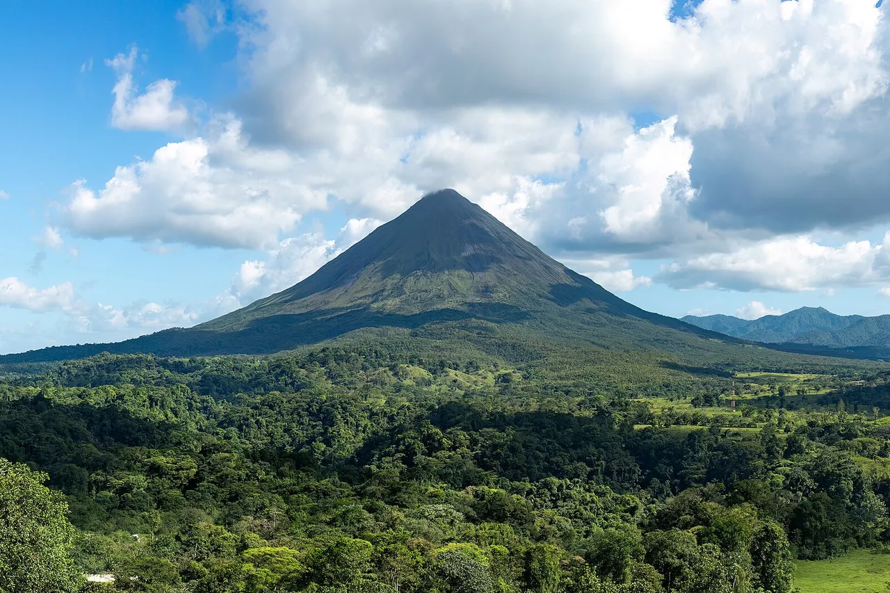 The cone of the Arenal volcano rises above a wide stretch of green rainforest under a blue sky with white clouds