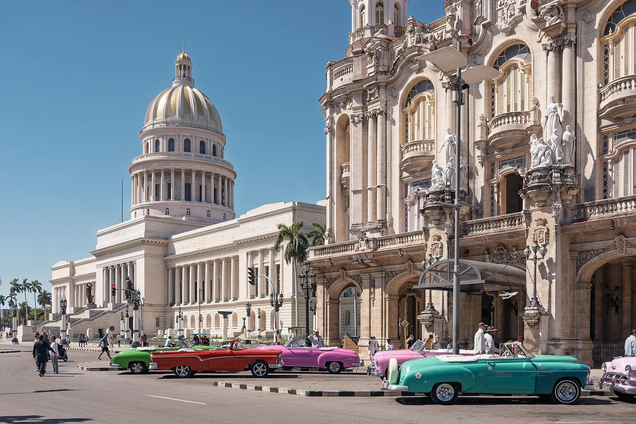 The domed National Capitol in Havana under a blue sky, with brightly painted classic American cars parked in front of the ornate Gran Teatro