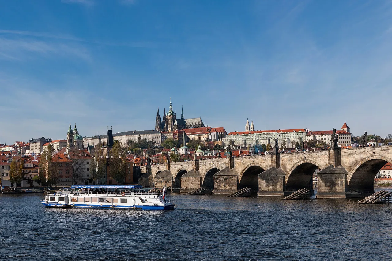 Prague Castle and St. Vitus Cathedral above the red roofs of the Lesser Town, with the stone arches of the Charles Bridge crossing the Vltava river