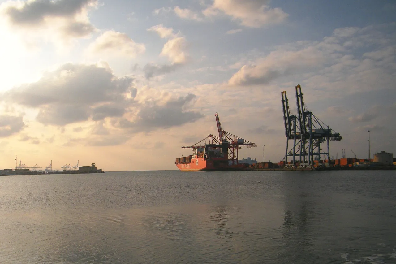 Container cranes and a red container ship at the Port of Djibouti, seen across calm water under a cloudy evening sky