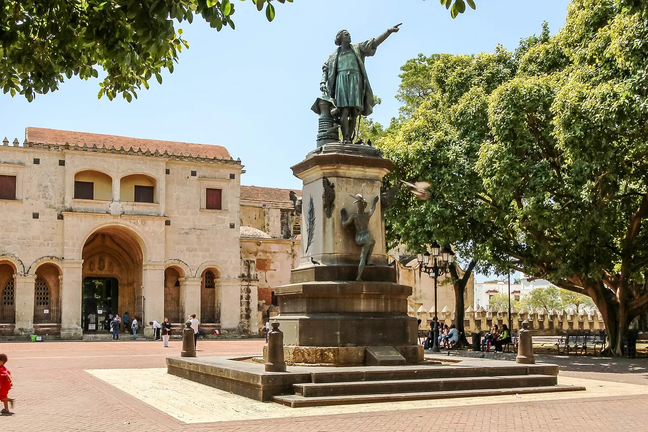 A bronze statue of Christopher Columbus on a tall stone pedestal stands in a sunny square shaded by trees, in front of the old stone cathedral of Santo Domingo