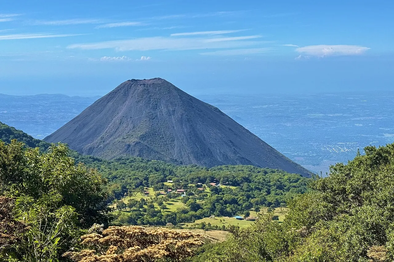The bare gray cone of the Izalco volcano rises from green forest, with farmland and the Pacific coast in the distance