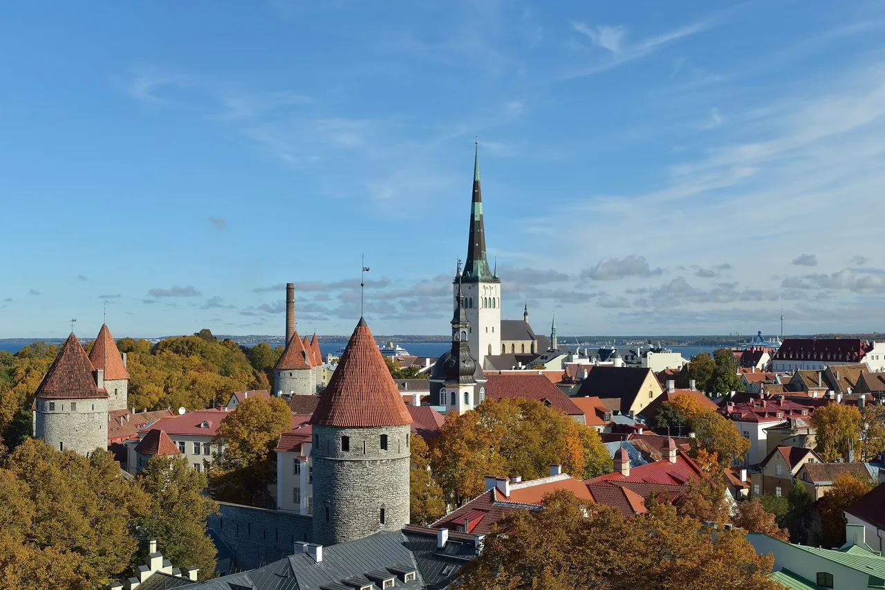Round stone towers with red cone roofs and the tall green spire of St. Olaf's Church above the old town of Tallinn, with autumn trees and the sea behind