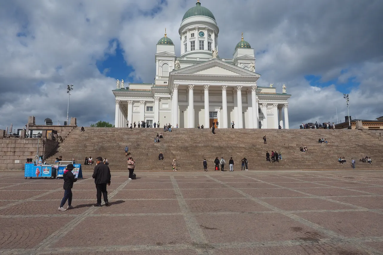 The white Helsinki Cathedral with its green domes stands above wide granite steps on Senate Square, under a cloudy sky