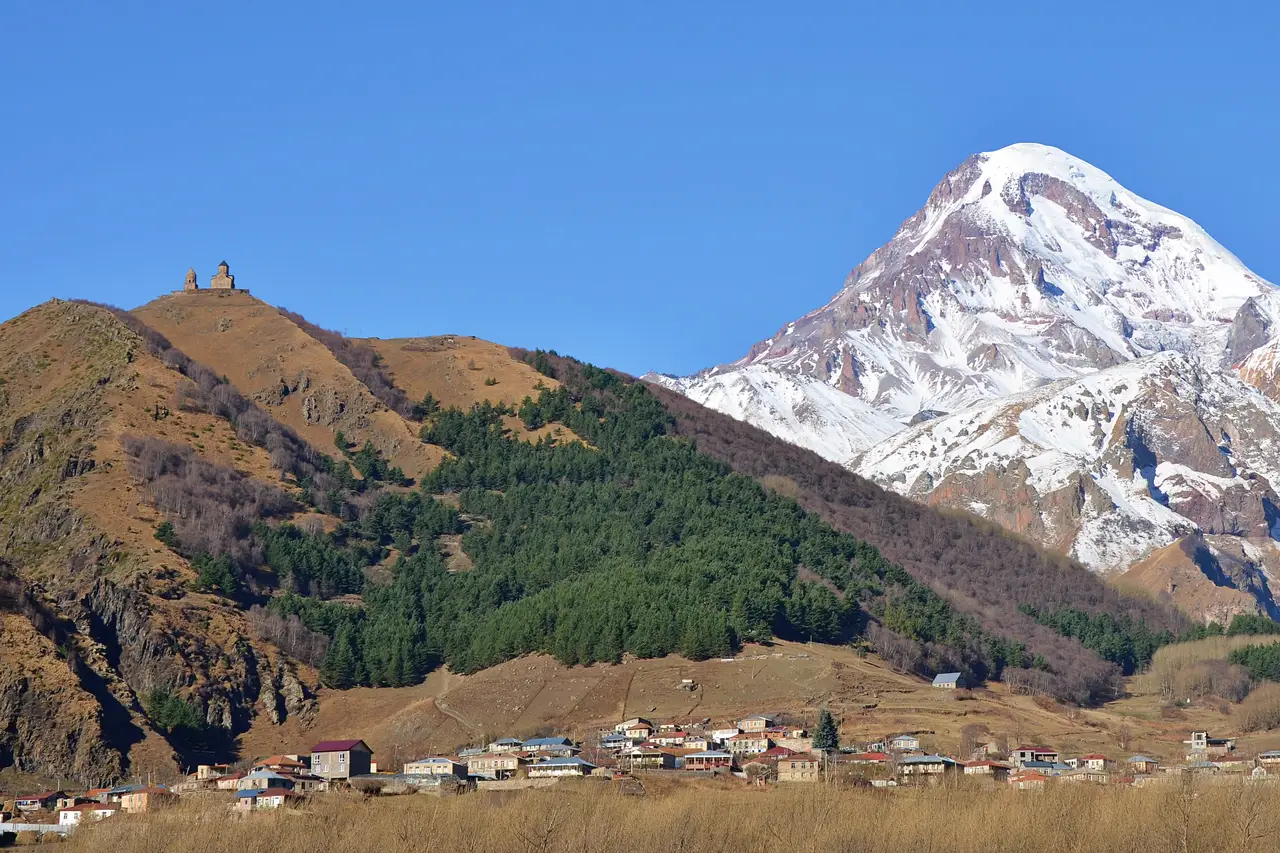 A small stone church on a brown hilltop above the village of Stepantsminda, with the snow-covered peak of Mount Kazbek under a clear blue sky