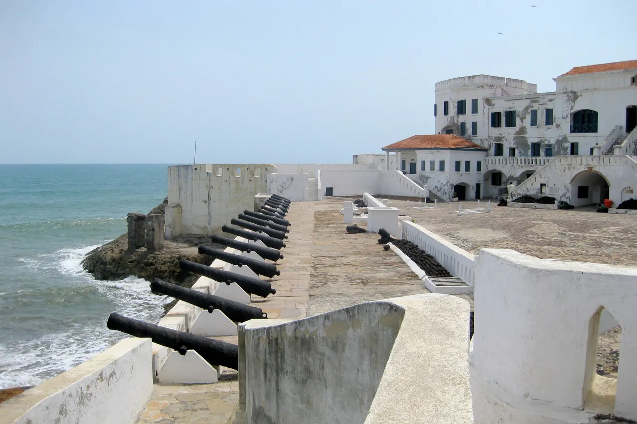 Old black cannons line a white fortress wall above the sea, with a whitewashed castle building behind
