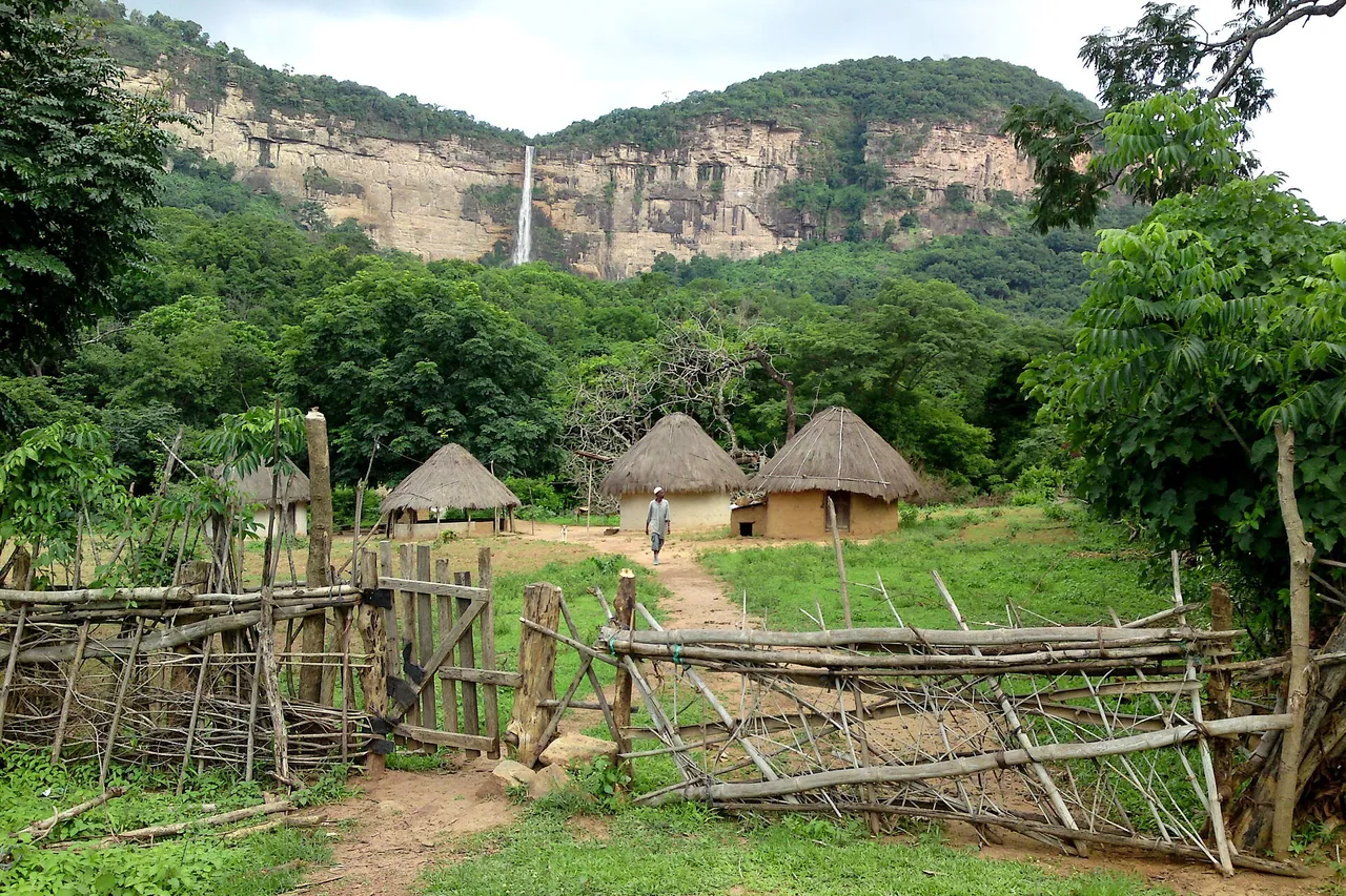 Round thatched huts behind a wooden fence in a green valley, with a thin waterfall falling from a long sandstone cliff behind them