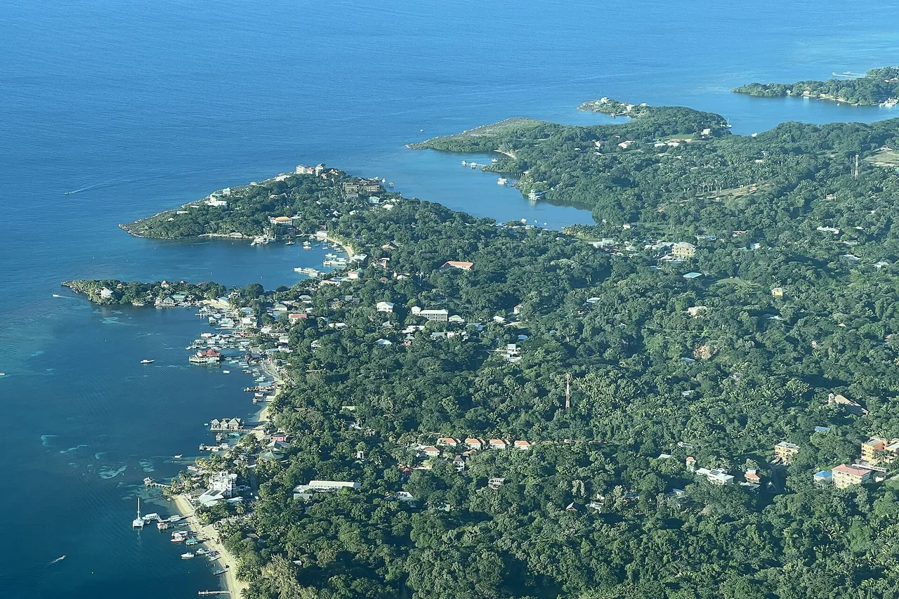 Aerial view of the green, wooded western tip of Roatán island, with the houses and docks of West End along a turquoise Caribbean shore