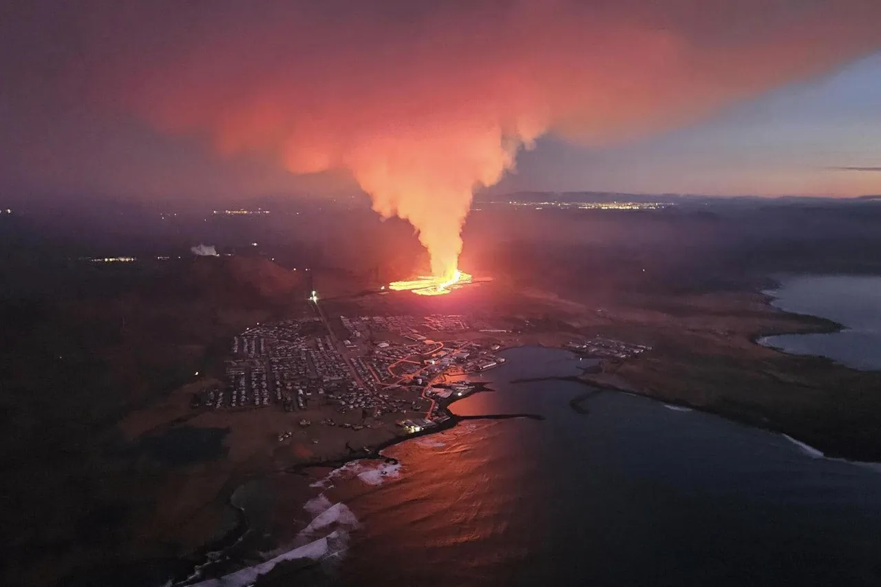 The town of Grindavík at dusk from the air, with a glowing lava eruption and a tall plume of smoke just beyond the houses