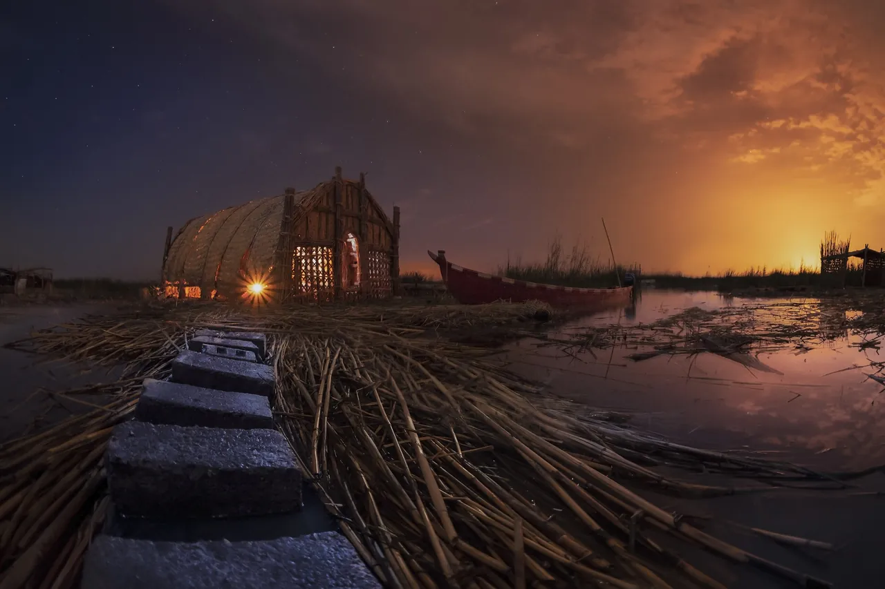 A lit reed guesthouse, a mudhif, and a canoe on the water of the Mesopotamian Marshes at dusk